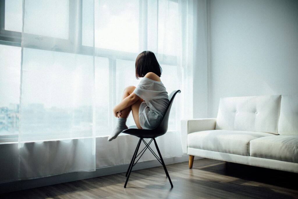 うつ病 用語集 1 うつ病の女性が椅子の上で膝を抱えて外を眺めている